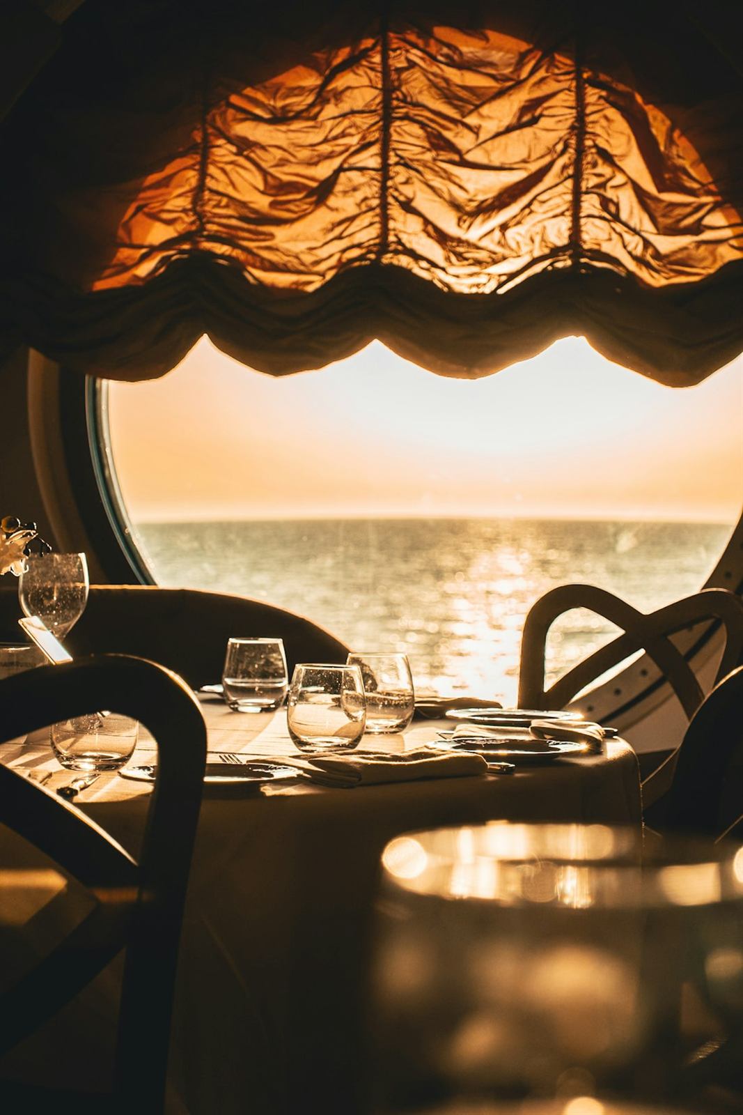 Gedekte tafel in een scheepsrestaurant, met zonsondergang op zee door een groot rond raam